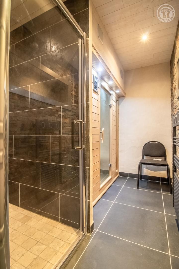 A bathroom with a glass shower enclosure, a tiled floor, and a chair.
