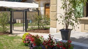 A courtyard with a grassy area, colorful flower beds, a potted plant, and a building with a stone facade and green shutters. - LA Cure Gourmande - LA Sure (SAINT CHRISTOPHE LA GROTTE)