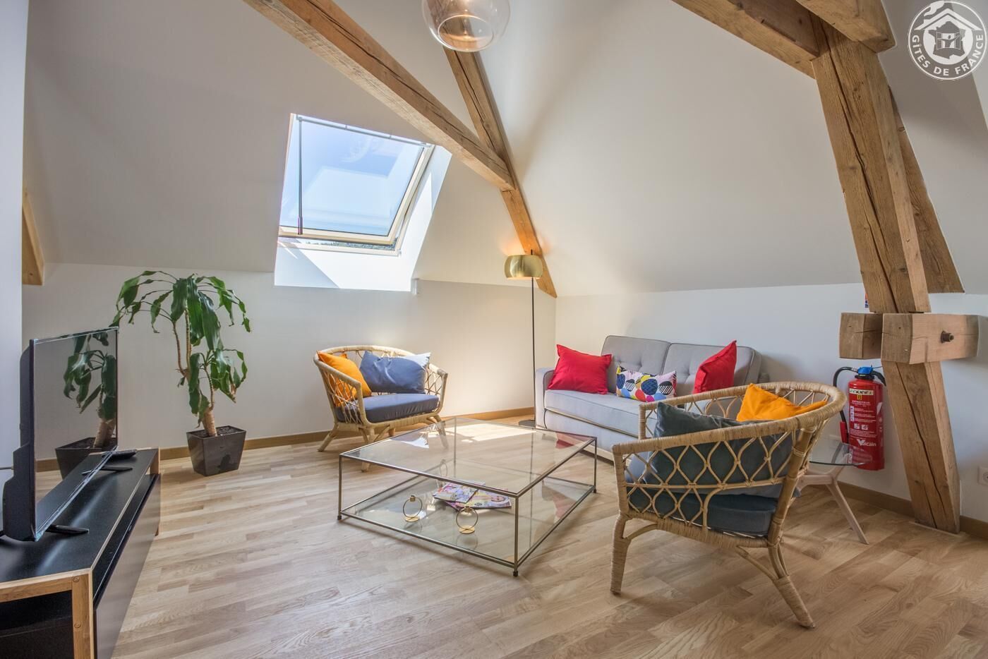 Ein gemütliches Loft mit schrägem Dach, einem Oberlicht, einem Glastisch, Korbmöbeln und einem Feuerlöscher.