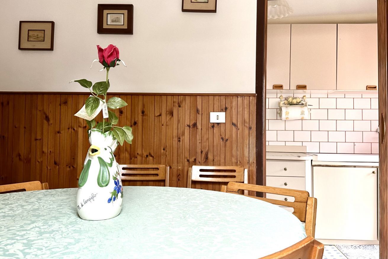 Una mesa de comedor con un florero, paneles de madera en la pared y una cocina al fondo.