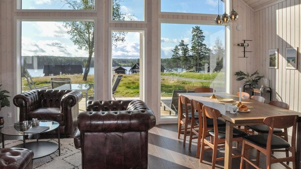 A spacious room with large windows offering a view of a lake and forest. There is a dining area with wooden furniture and a leather sofa. - 2 bedroom cozy home in årjäng (Årjäng)