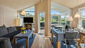 Un espacio habitable moderno y de planta abierta con interior de madera, televisor de pantalla plana, zona de comedor con mesa y sillas, y ventanas grandes que ofrecen vistas al exterior.