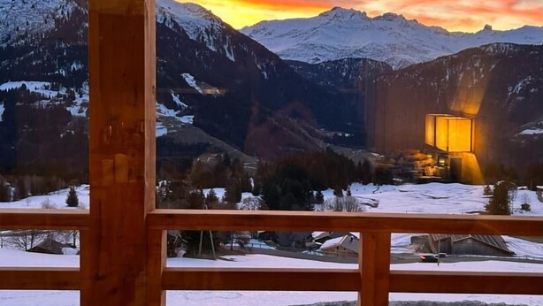 Un balcon en bois offrant une vue sur des montagnes enneigées et un coucher de soleil.