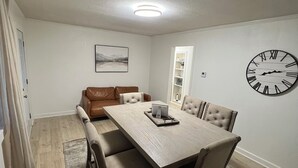 A dining area with a wooden table, cushioned chairs, a clock on the wall, and a framed picture above the table. - Cozy home in a peaceful country setting with stocked pond. Relax and unwind. (Bridgeport)