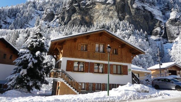 Un chalé de madera con una escalera que lleva a la entrada, rodeado de árboles nevados y un paisaje de montaña.