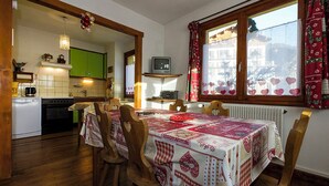 Un comedor con una mesa dispuesta para cuatro, un televisor en un estante y una ventana con cortinas estampadas con corazones.