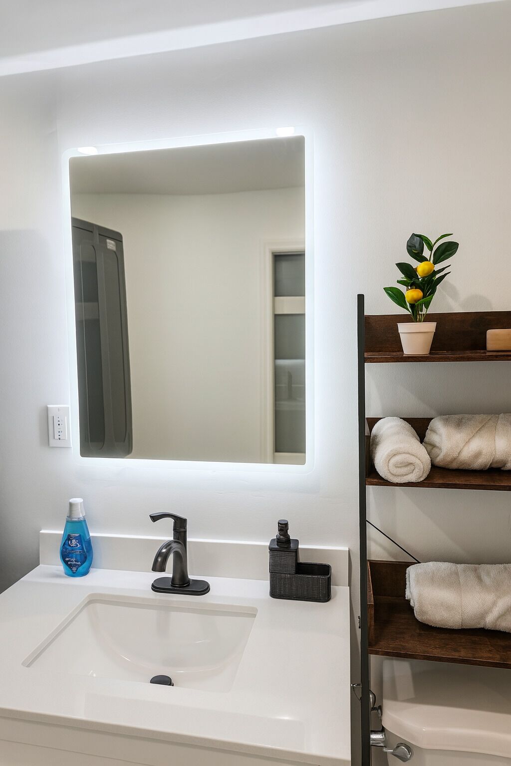 A bathroom with a white sink, a mirror with a light, a potted plant, and a shelf with towels.