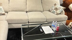 Un salon avec un canapé beige, une table basse en verre sur laquelle se trouvent une télécommande, des bouteilles d’eau et une canette de soda.