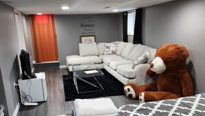 Une chambre d’hôtel moderne avec un canapé d’angle, un grand ours en peluche, un téléviseur à écran plat et un lit avec une literie à motifs.