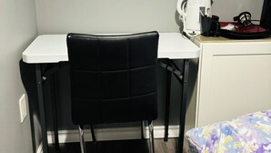 Une petite pièce avec un bureau, une chaise noire, un lit recouvert d’une courtepointe à motifs floraux, une bouilloire blanche et un sac noir posé sur le bureau.
