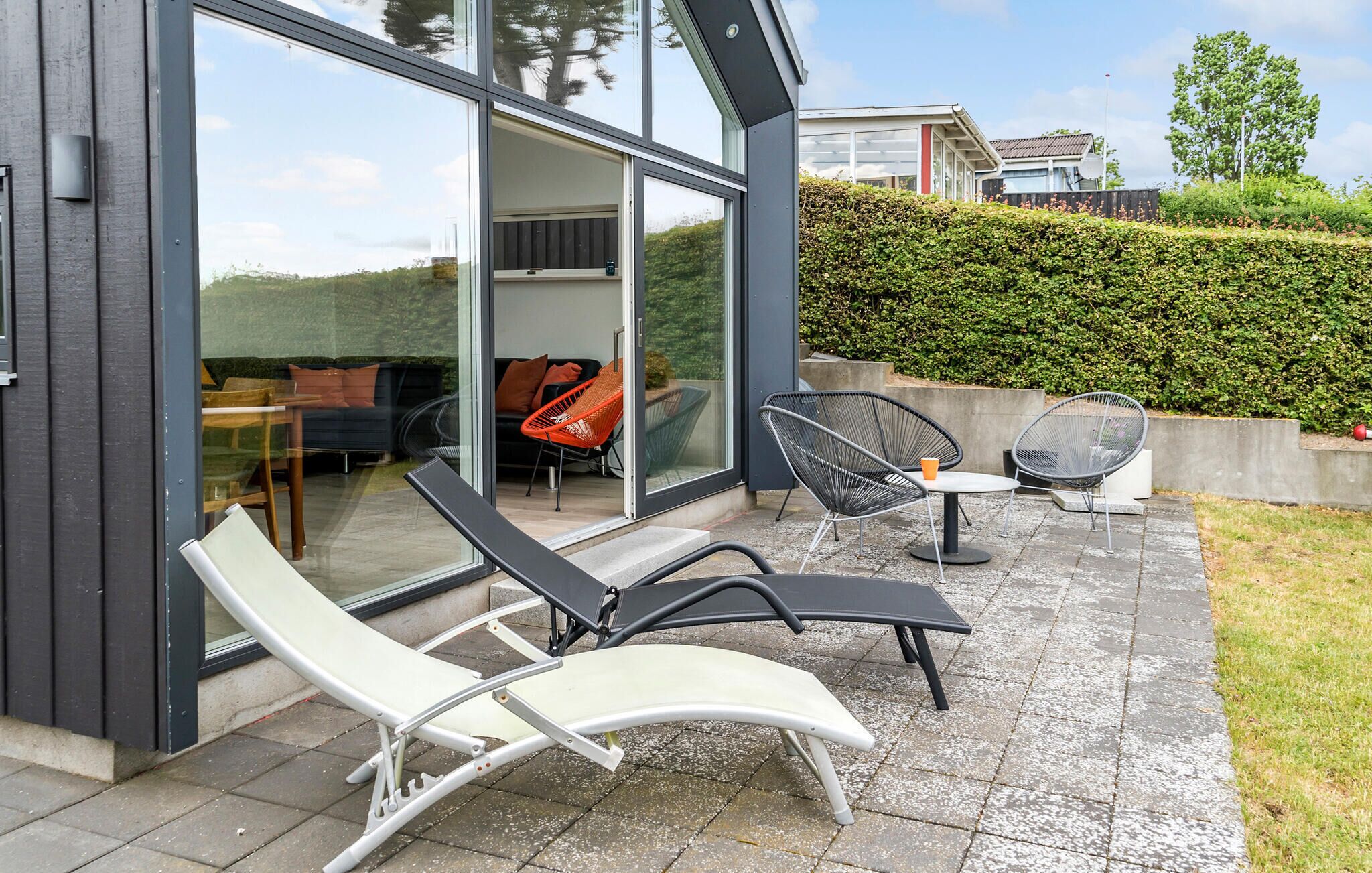 A modern outdoor patio with lounge chairs and a table, large glass doors leading to an interior space, and a well-maintained hedge in the background.