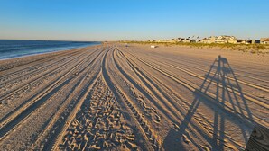 A wide sandy beach with tire tracks leading to the water, a clear sky, and a row of houses in the background.