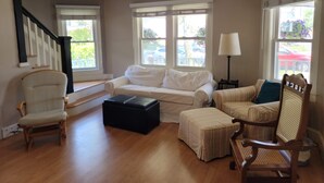 A living room with a sofa, armchairs, a wooden chair, a black ottoman, and a lamp.