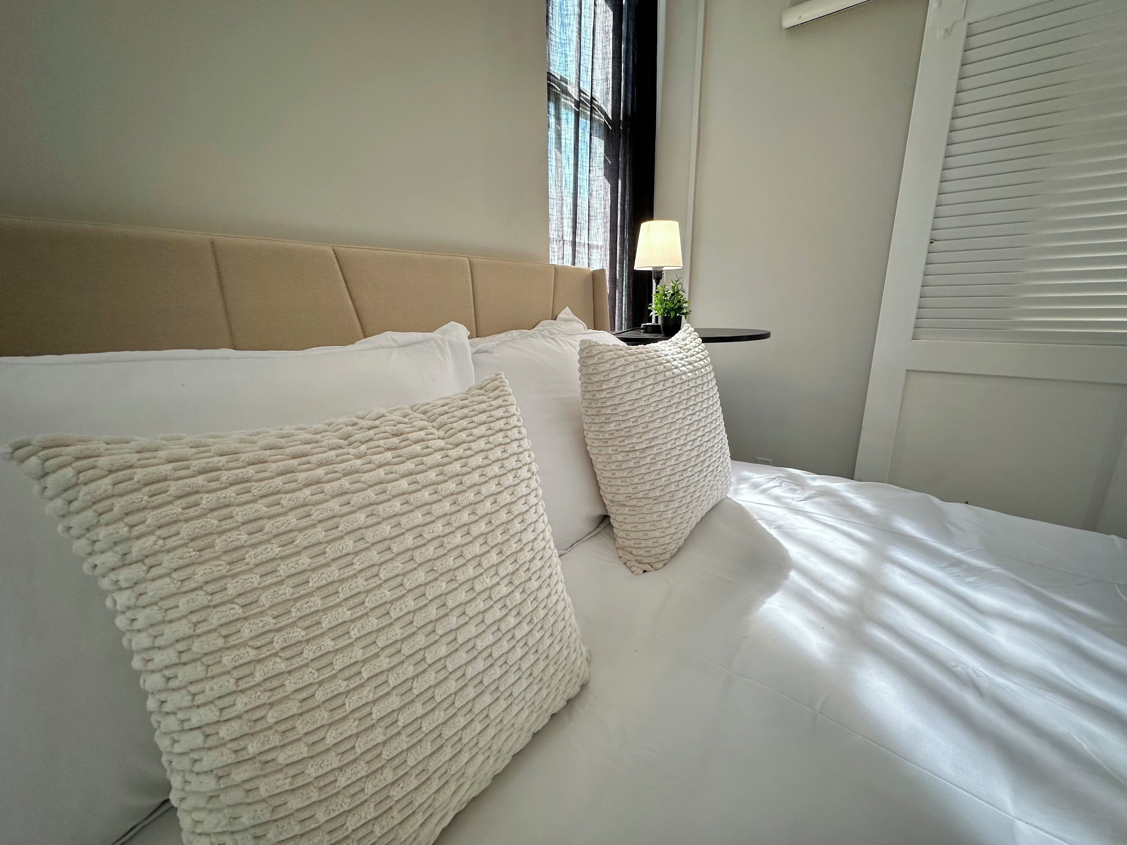 Un lit avec une literie blanche et deux coussins à motifs, une table de chevet avec une lampe et une fenêtre avec des rideaux.