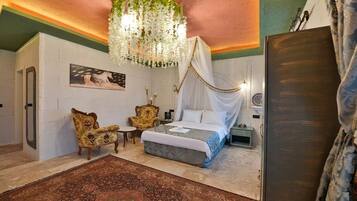 A bedroom with a bed, a chair, a table, a hanging floral chandelier, and a painting on the wall.