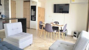 A modern living room with grey sofas, a dining area with a wooden table and chairs, a kitchen with a built-in oven, and a mounted TV.