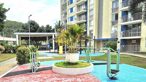 An outdoor gym area with exercise equipment, surrounded by a pool and greenery, in front of a multi-story building.