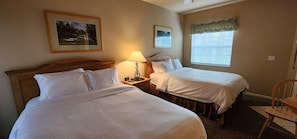 A hotel room with two single beds, a bedside table with a lamp, a chair, and a window with blinds.