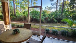 Una mesa de madera redonda con sillas, una planta en maceta y vistas a un jardín exuberante a través de grandes ventanales.