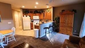 Una cocina con armarios de madera, un refrigerador blanco, un microondas y un lavamanos. Hay una mesa de comedor con sillas y un sofá marrón en la sala de estar.