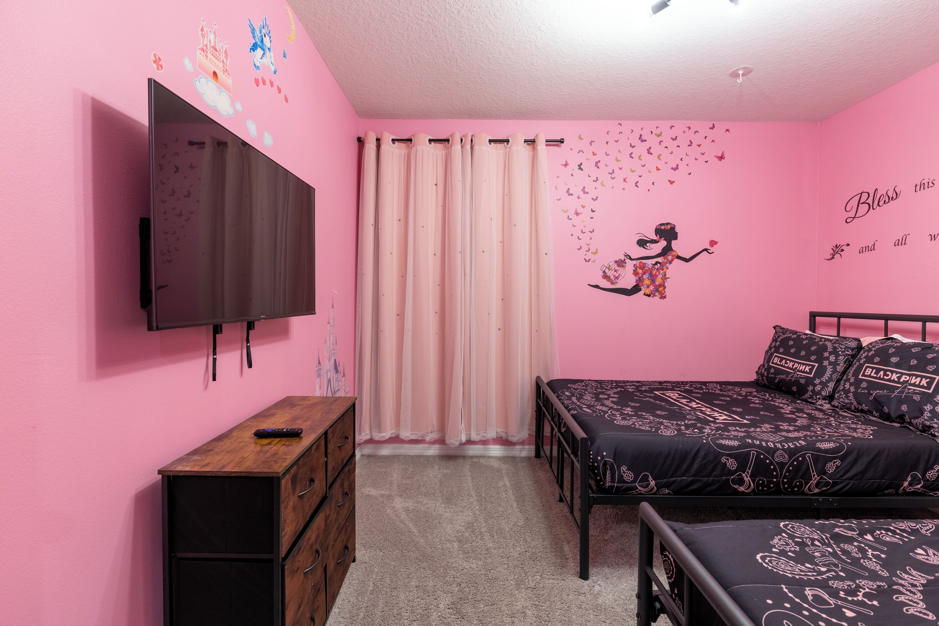 Une chambre à coucher décorée dans des tons roses, avec une télévision, une commode en bois, un lit avec une literie à motifs et un autocollant mural.