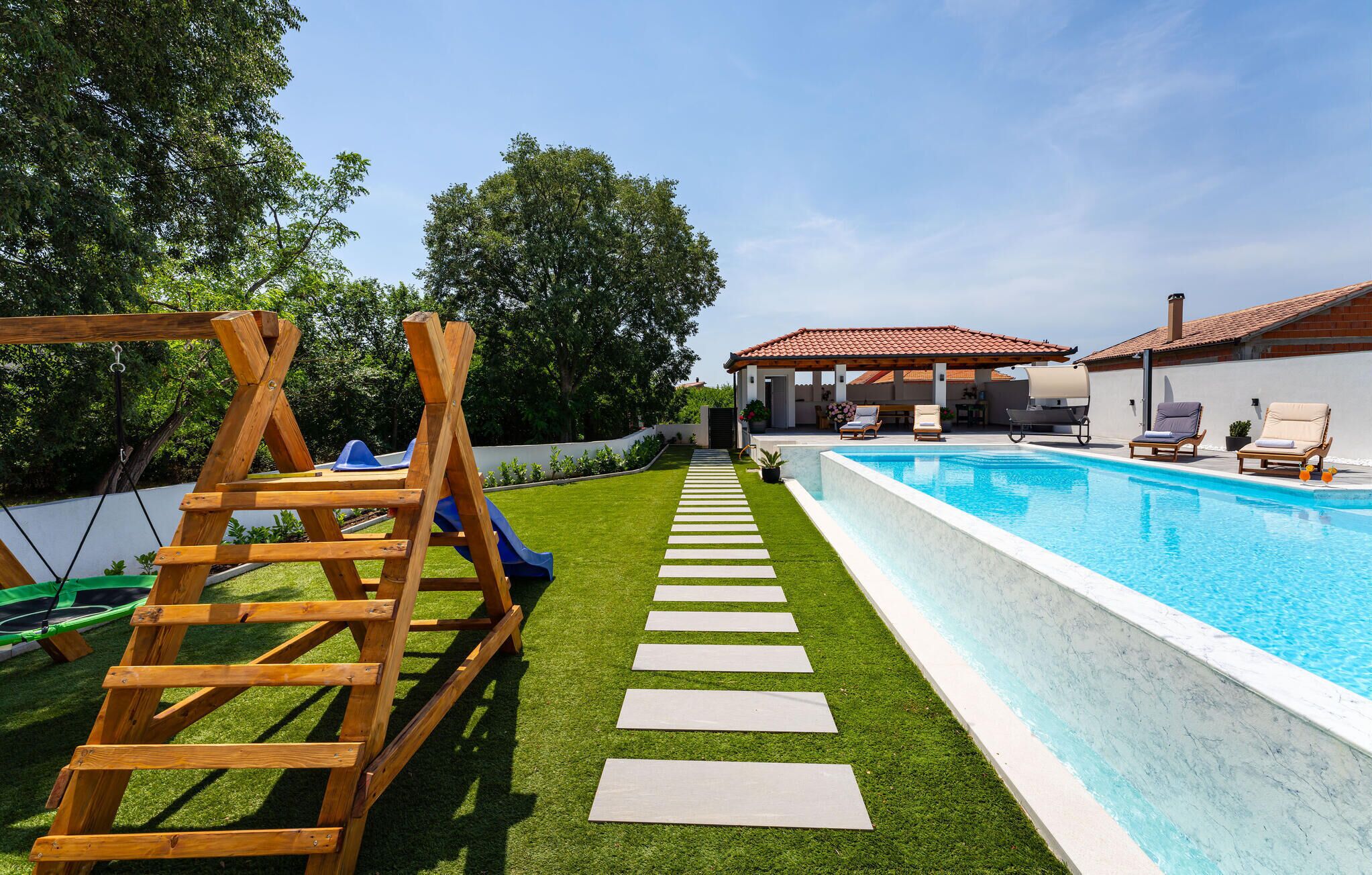 A backyard with a swimming pool, a wooden swing set, and a patio area with outdoor furniture.