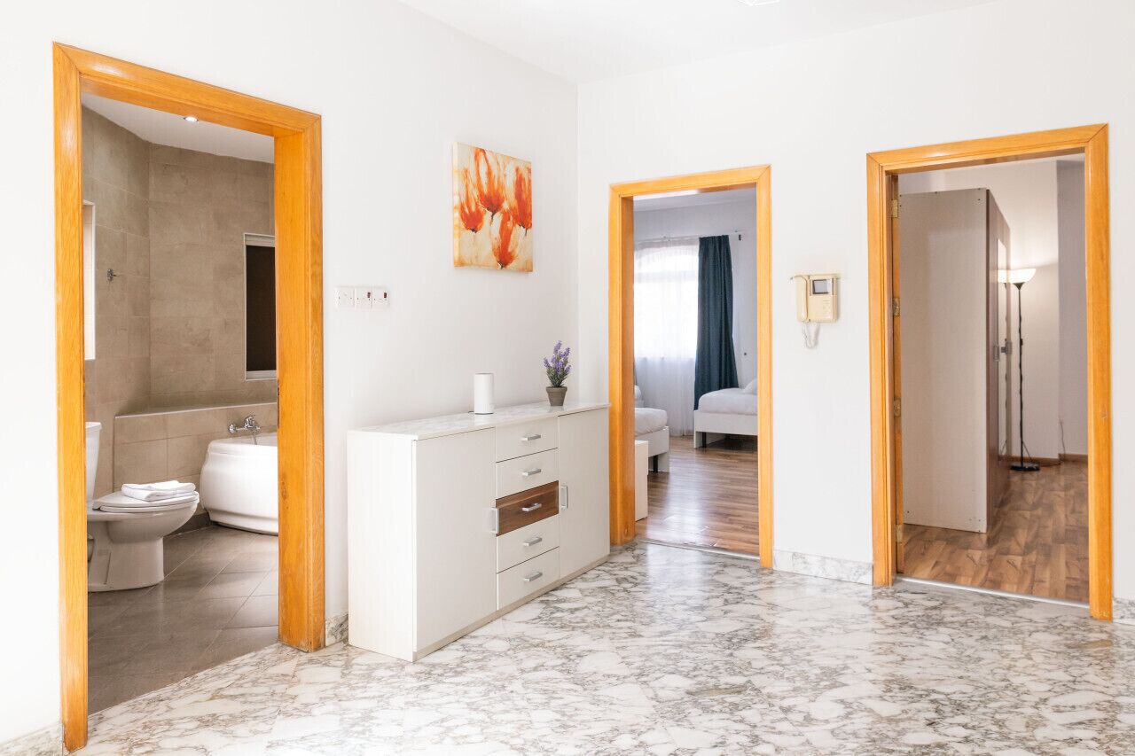 Un baño moderno con ducha, inodoro y lavamanos. Se ve un pasillo que conduce a un dormitorio con cama y mesita de noche.