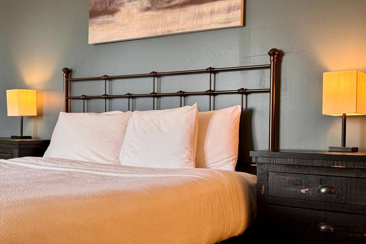 Una cama bien tendida con sábanas blancas, un cabecero de metal oscuro y una mesita de noche de madera con una lámpara.