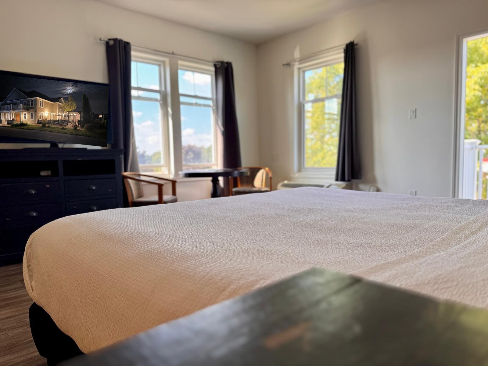Une chambre d’hôtel avec un grand lit, un téléviseur posé sur une commode, une table à manger et deux fenêtres avec des rideaux.