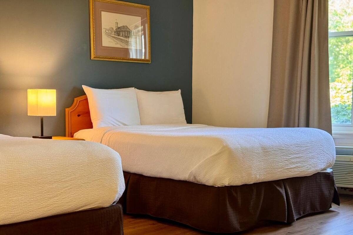 Habitación de hotel con dos camas, una mesita de noche con lámpara, un cuadro enmarcado en la pared y una ventana con cortinas.