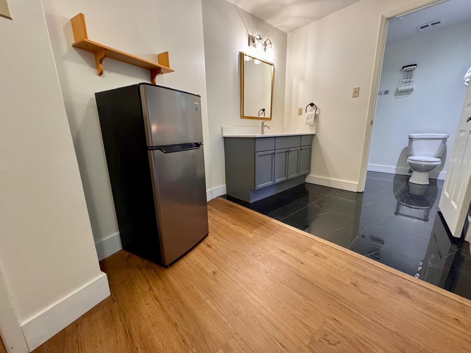 Une cuisine compacte avec un réfrigérateur noir, des étagères en bois, un miroir et des toilettes visibles à travers une porte en verre.