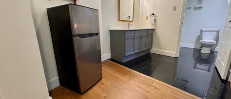 Une cuisine compacte avec un réfrigérateur noir, des étagères en bois, un miroir et des toilettes visibles à travers une porte en verre.