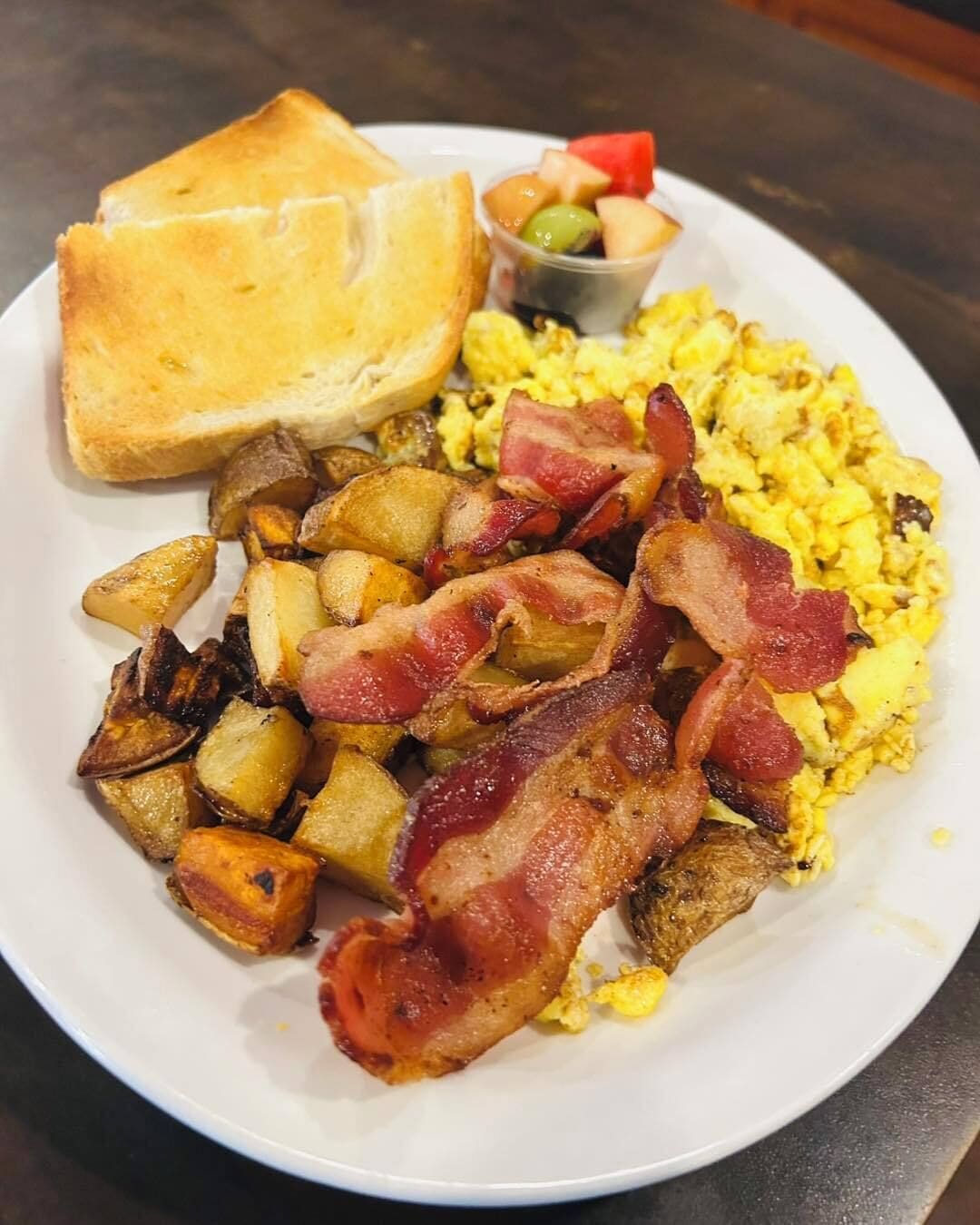 Un plato con tostadas, huevos revueltos, tocino, papas y una pequeña ensalada de frutas.