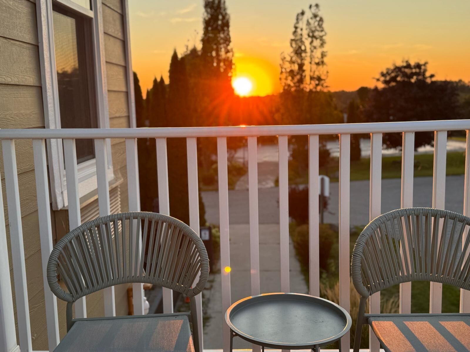 A balcony with two chairs and a small table, overlooking a sunset with the sun setting behind trees.