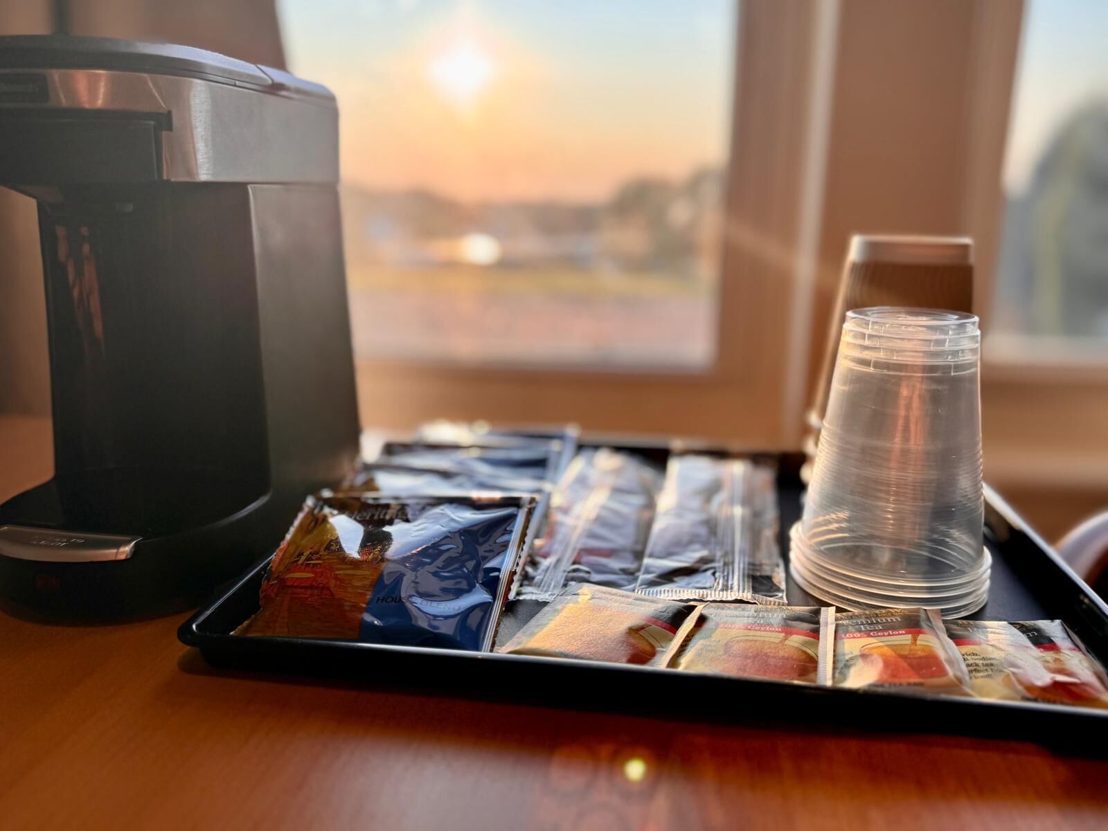 A tray with a coffee maker, a stack of clear plastic cups, and a magazine on a wooden table near a window with a view of the sunset.