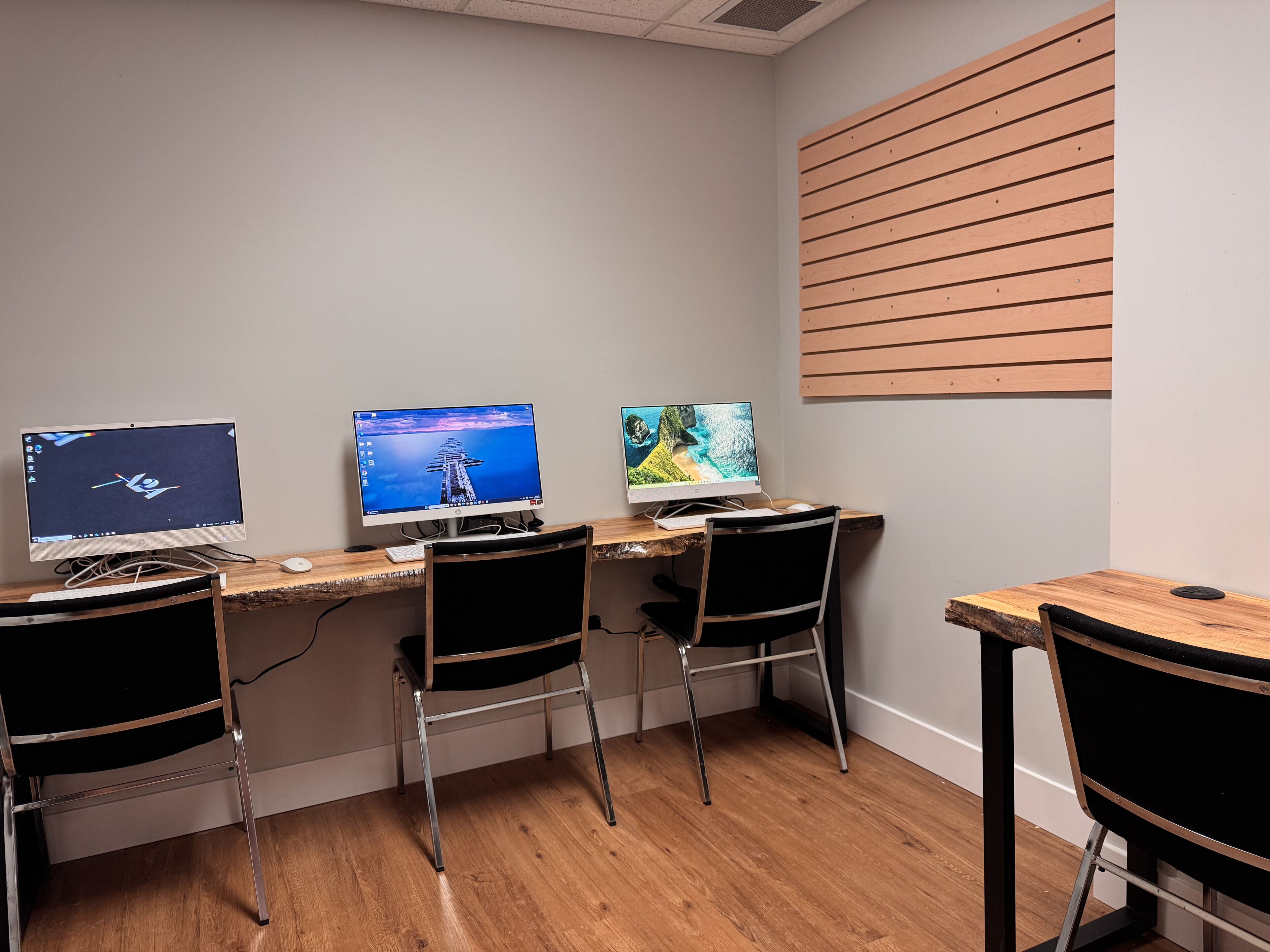 Un espacio de trabajo con tres escritorios, cada uno con un monitor de computadora, teclado y ratón. La habitación tiene suelo de madera y un techo inclinado.