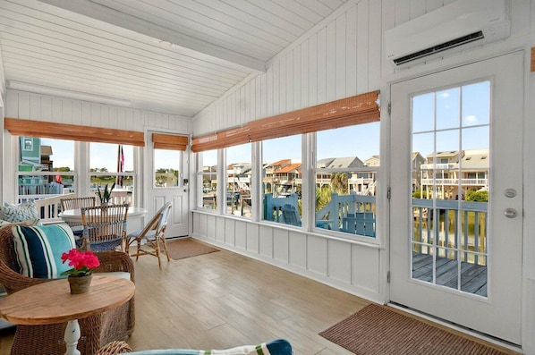 A sunroom with white paneled walls, large windows, wicker furniture, and a view of neighboring houses.
