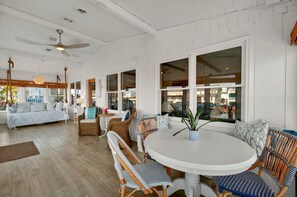 A spacious living room with a large round table, wicker chairs, and a sofa. There are large windows providing natural light and views of the outside.