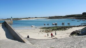 Paisaje costero con un faro, barcos y gente en una playa arenosa.