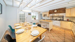 Une cuisine lumineuse avec une table en bois dressée pour le repas, un luminaire suspendu et un mur en briques orné d’ustensiles de cuisine.
