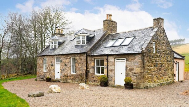 Une maison en pierre avec une allée gravillonnée, un petit jardin et une vue sur la campagne environnante.