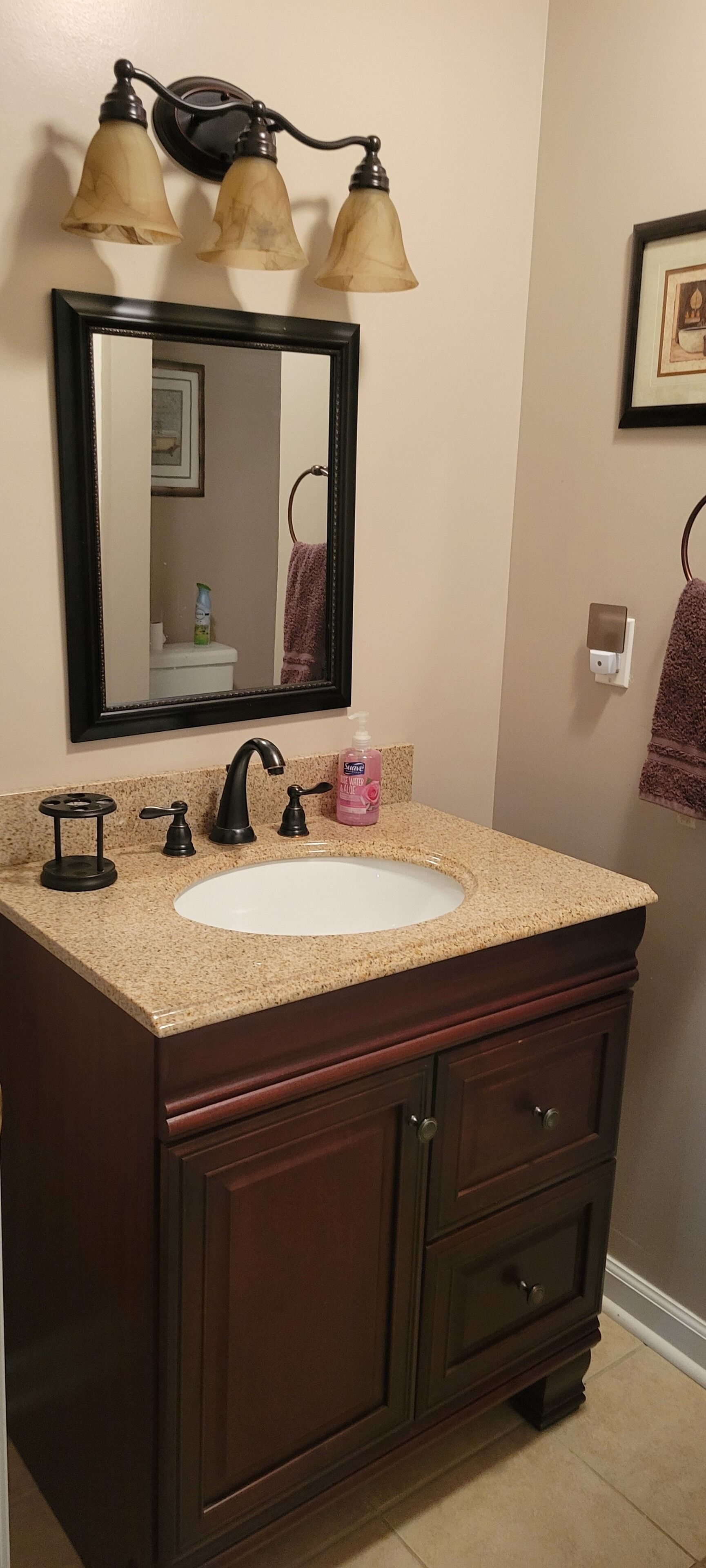 A bathroom with a beige granite countertop, a white sink, a dark wood vanity, and a black-framed mirror.