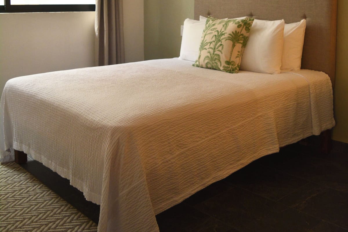 A neatly made bed with a headboard, a window with curtains, and a patterned rug on the floor.
