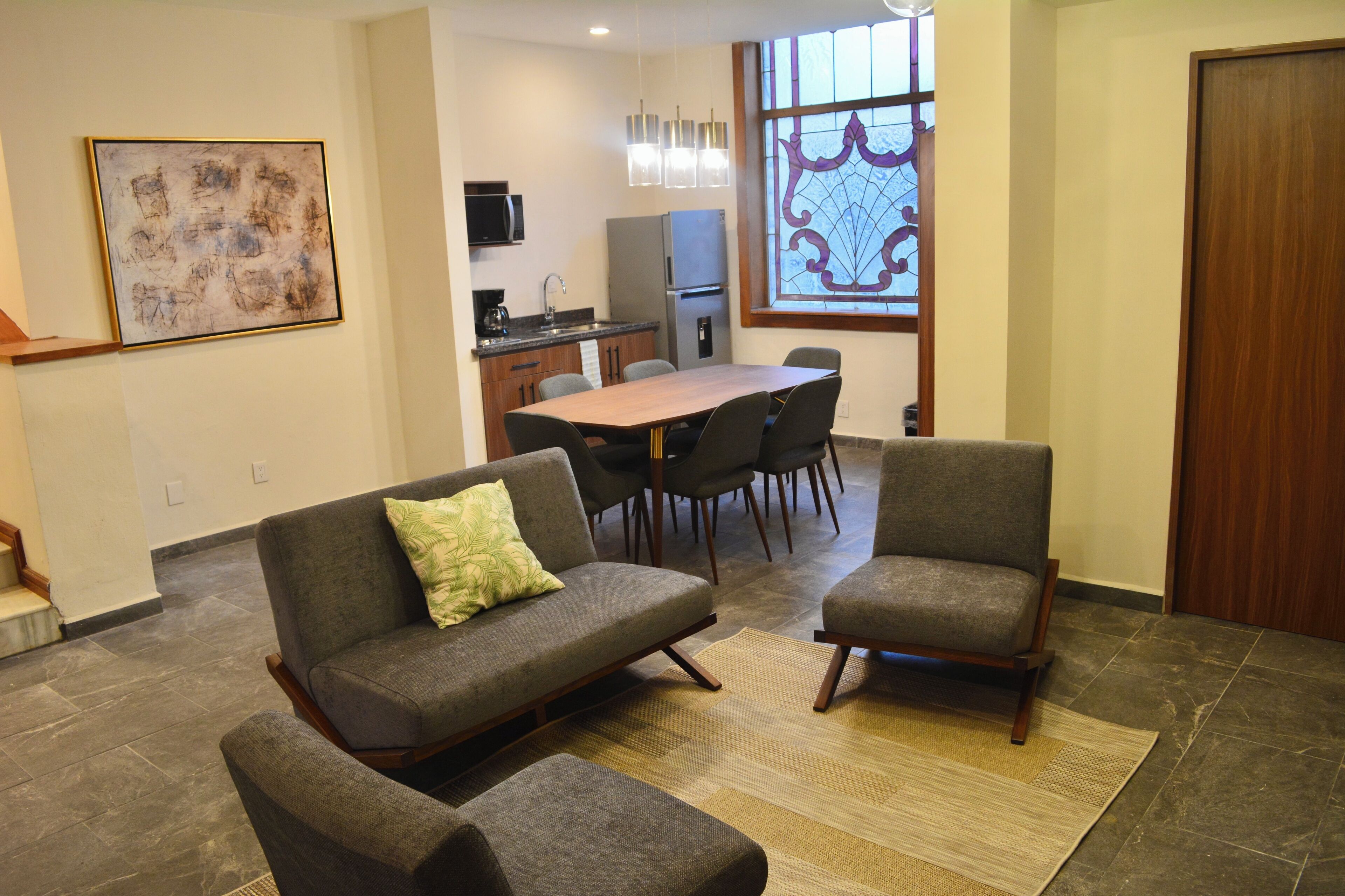 A modern living room with a sofa, armchair, and a dining table with chairs. There is a kitchen area with appliances and a window with decorative glass.