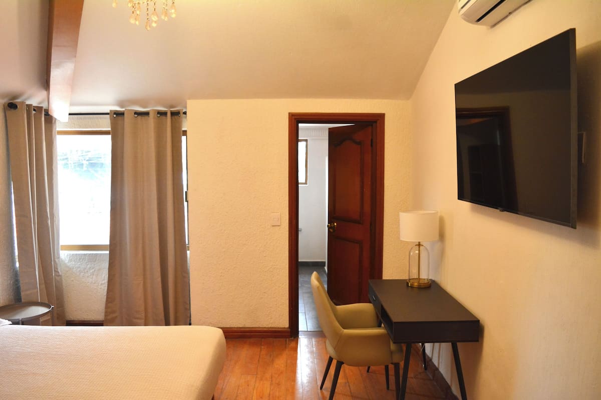 A hotel room with a bed, a desk, a chair, a lamp, and a television on the wall.