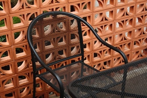 Una silla de metal con respaldo de malla y una mesa de metal, frente a un muro de ladrillo con un diseño geométrico.