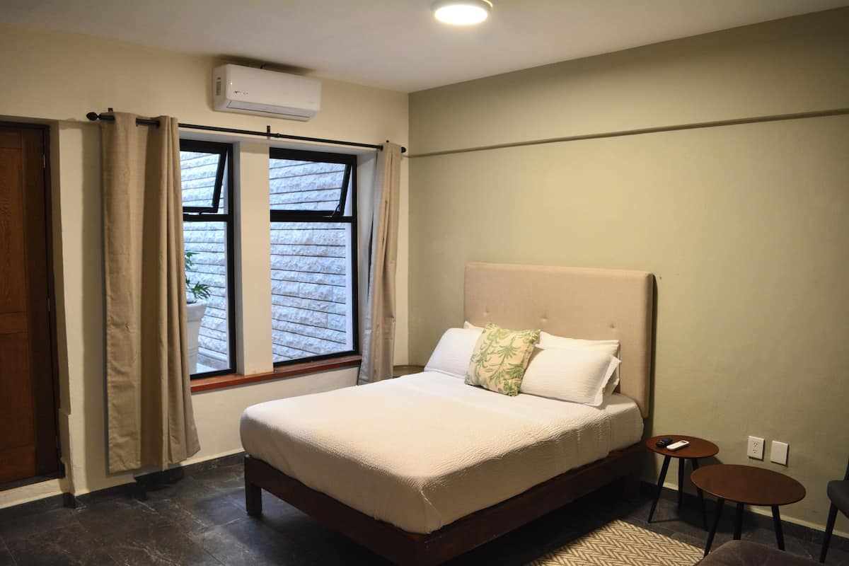 A bedroom with a large bed, a window with curtains, a small table, and a wall-mounted air conditioner.