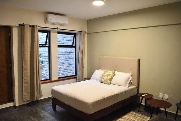 A bedroom with a large bed, a window with curtains, a small table, and a wall-mounted air conditioner. - Casa Palmas (Zapopan)