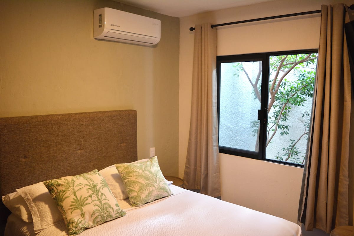A hotel room with a bed, a window with curtains, and an air conditioning unit on the wall.