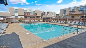 Une piscine avec des chaises longues et une clôture en bois, entourée d’immeubles à plusieurs étages dotés de balcons.